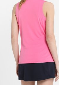 Kobieta w bezrękawniku w kolorze różowym i ciemnoniebieskiej spódnicy, pokazana od tyłu na jednolitym tle.