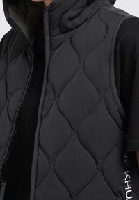 Close-up of a person wearing a black quilted sleeveless vest over a black long-sleeve shirt with a zipper partially open.