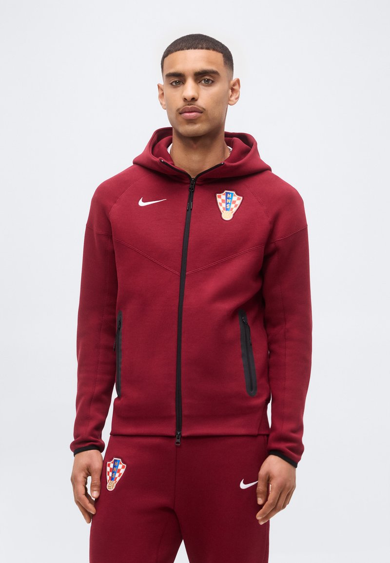 Νεαρός άνδρας φορώντας μαρόν φόρμα Nike με τα εμβλήματα της κροατικής ποδοσφαιρικής ομάδας, στέκεται μπροστά σε απλό ανοιχτό φόντο.