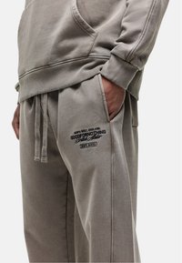 Hellegrauer Hoodie mit einer Vorderseite Tasche und gerippten Bündchen, kombiniert mit passenden Jogginghosen, die ein gesticktes Logo in Schwarz tragen.