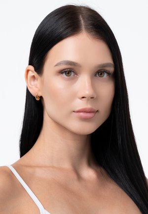 Goldene Creolen mit einem eleganten, geschwungenen Design. Das Modell hat lange schwarze Haare und einen glatten Teint und trägt ein einfaches weißes Oberteil.