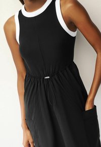 Schwarzes ärmelloses Kleid mit gerippter Textur, weißen Rundhalsausschnitt, elastischem Taillenband und seitlichen Taschen.