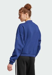 Sudadera azul con un corte holgado, puños acanalados y un escote alto. Presenta un sutil detalle del logo en la manga. Combinada con pantalones negros.