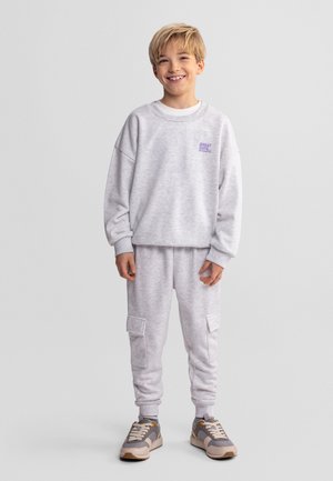 Uśmiechnięty chłopiec z blond włosami stoi w jasnoszarej bluzie i dopasowanych spodniach cargo typu joggery, nosząc szaro-beżowe tenisówki na jednolitym tle.