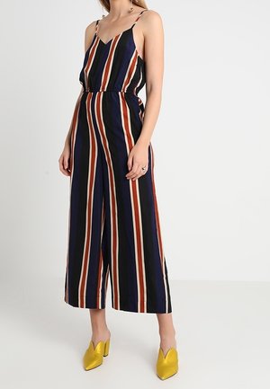 Kombinezon w paski na cienkich ramiączkach, z pionowymi paskami w kolorze granatowym, czerwonym i kremowym. Szerokie nogawki i elastyczna talia. Żółte buty wsuwane.