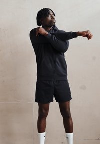 Sort zip-up hættetrøje og shorts lavet af glat stof. Modellen står i en strækpose, iført hvide sportsokker og sorte sko.