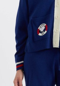 Marineblauwe gebreide cardigan met witte knopen, gestreepte manchetten en een patch met Snoopy in een cirkelvormig ontwerp. Zak aan de linkerkant.