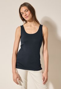 Pitkähiuksinen nainen, jolla on päällään istuva musta hihaton paita ja vaaleanbeiget housut, seisoo yksinkertaisen vaalean seinän edessä.