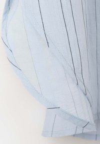 Tissu bleu clair avec de fines rayures verticales noires, montrant un gros plan détaillé d'un poignet de manche de chemise avec des coutures visibles.