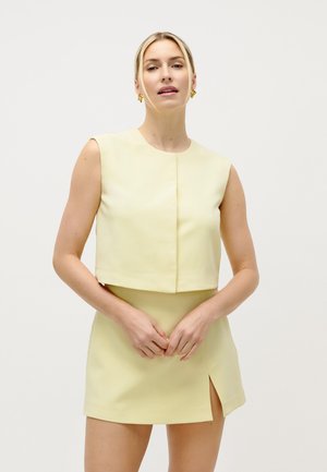 Ženska, oblečena v brezrokavnik bledo rumene kratke majice in ujemajočo se mini krilo s sprednjim razrezom, stoji pred enobarvnim belim ozadjem.