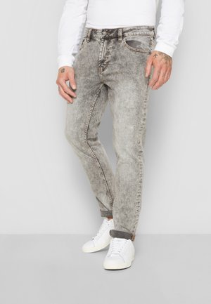 Homme portant un jean gris délavé à l'acide, les revers retroussés, des baskets blanches et une chemise blanche à manches longues, debout devant un fond uni.