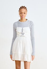 Vestido blanco de mezcla de lino con recorte frontal y detalle de lazo, superpuesto a una blusa de manga larga ribeteada en gris claro. Textura suave y corte relajado.