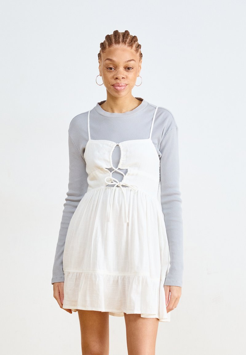 Vestido blanco de mezcla de lino con recorte frontal y detalle de lazo, superpuesto a una blusa de manga larga ribeteada en gris claro. Textura suave y corte relajado.