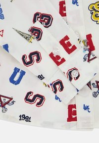 Poignet de chemise blanche avec des lettres de style varsity rouges, bleues et noires et de petites impressions de personnages de dessin animé, fixé avec deux boutons transparents.