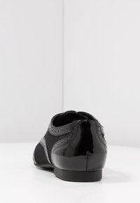 Chaussure noire avec un dos en cuir verni, un côté en tissu texturé, des détails en perforation et un talon bas. Design minimaliste aux lignes épurées.