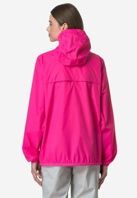 Donna che indossa una giacca a vento con cappuccio di colore rosa acceso, vista da dietro, con pantaloni chiari su uno sfondo neutro.