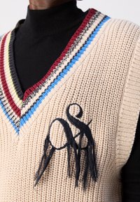 Homme portant un gilet en tricot beige à col en V rayé coloré sur un col roulé noir, avec un logo brodé noir orné de franges.