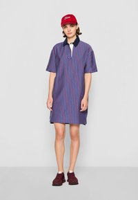 Vestido polo às riscas em azul e bordô, com mangas curtas, um colarinho azul-marinho e fendas laterais, combinado com uma boné vermelho e ténis bordô.