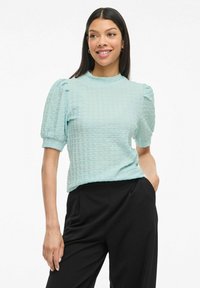Top azzurro pallido con texture, maniche a sbuffo corte e colletto alto, abbinato a pantaloni neri, che mette in evidenza un design strutturato ma morbido.