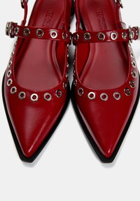 Rote Kunstleder-Ballerinas mit spitzen Zehen, silbernen Ösenakzenten und verstellbaren Riemen am Knöchel. Flache schwarze Sohlen runden das Design ab.