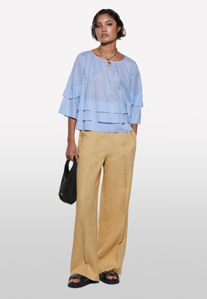 Blouse en légère superposition bleu clair à manches larges, associée à un pantalon en lin beige ample. Un sac tissé noir et des sandales plates noires complètent le look.