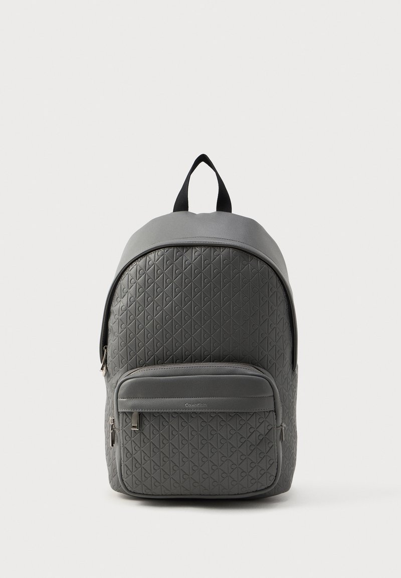 Grå rygsæk lavet af tekstureret syntetisk materiale, med et trekantet mønster, en frontlomme med lynlås og et bærehåndtag på toppen.