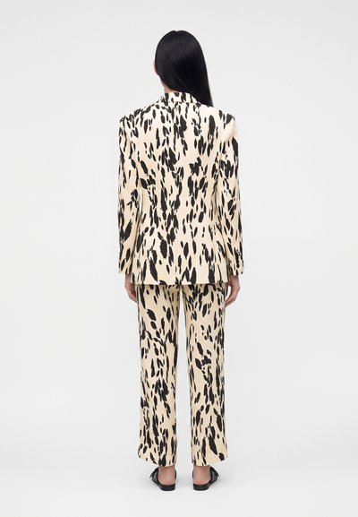 Femme aux longs cheveux noirs, vue de dos, portant un costume couleur crème avec des taches noires irrégulières et des chaussures plates noires.