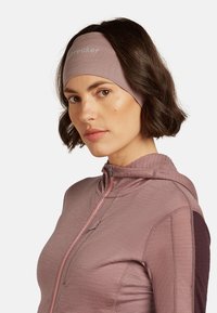 Fascia per capelli di un leggero viola, realizzata in materiale elastico con una texture liscia, caratterizzata da un logo e una vestibilità aderente. Il modello indossa una giacca con zip.