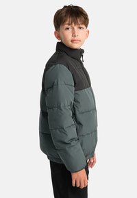 Ragazzino che indossa un giubbotto imbottito bicolore con la sezione superiore nera e la sezione inferiore grigio-verde, in piedi di lato su uno sfondo bianco.