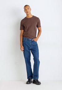 Tricou din bumbac maro asociat cu blugi de denim negru. Modelul poartă pantofi negri. Design simplu, croială lejeră, fără modele sau logo-uri vizibile.