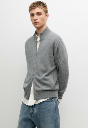 Jeune homme portant un pull gris zippé sur une chemise blanche et un jean bleu ample, debout devant un fond clair uni.