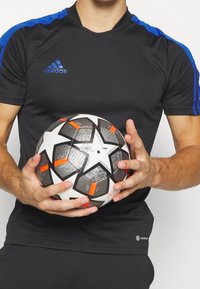 Jersey atlético negro con franjas azules en los hombros y escote redondo. El jugador sostiene un balón de fútbol negro y blanco con acentos en naranja y un diseño de estrella.