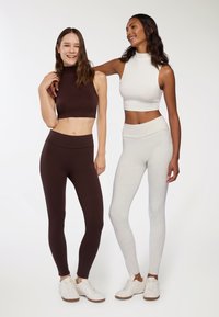 Due modelle indossano top aderenti e leggings. Una modella in marrone scuro e l'altra in grigio chiaro, entrambe con sneakers bianche.