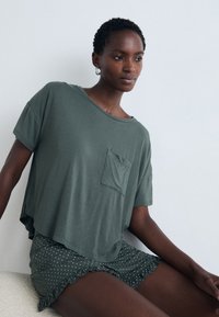 Kvinna som bär en lös grön t-shirt och gröna prickiga shorts med volangkant, sittande mot en ljusgrå bakgrund.