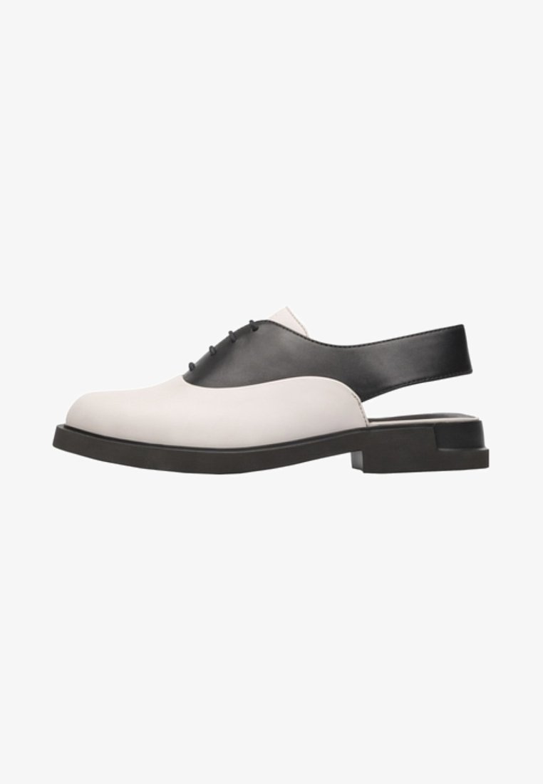 Chaussure à bride arrière en cuir noir et blanc avec un bout arrondi, une semelle plate et un talon découvert. Présente un détail discret de lacets sur le devant.
