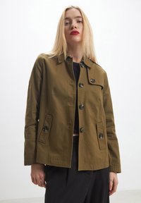 Donna bionda che indossa una giacca verde oliva con bottoni sopra una maglietta e pantaloni neri, in piedi contro uno sfondo bianco semplice.