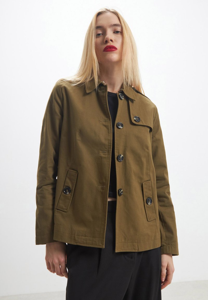 Donna bionda che indossa una giacca verde oliva con bottoni sopra una maglietta e pantaloni neri, in piedi contro uno sfondo bianco semplice.