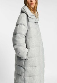 Abrigo tipo puffer gris claro con cuello alto, diseño acolchado y costuras prominentes. Cuenta con cierres a presión y bolsillos laterales.