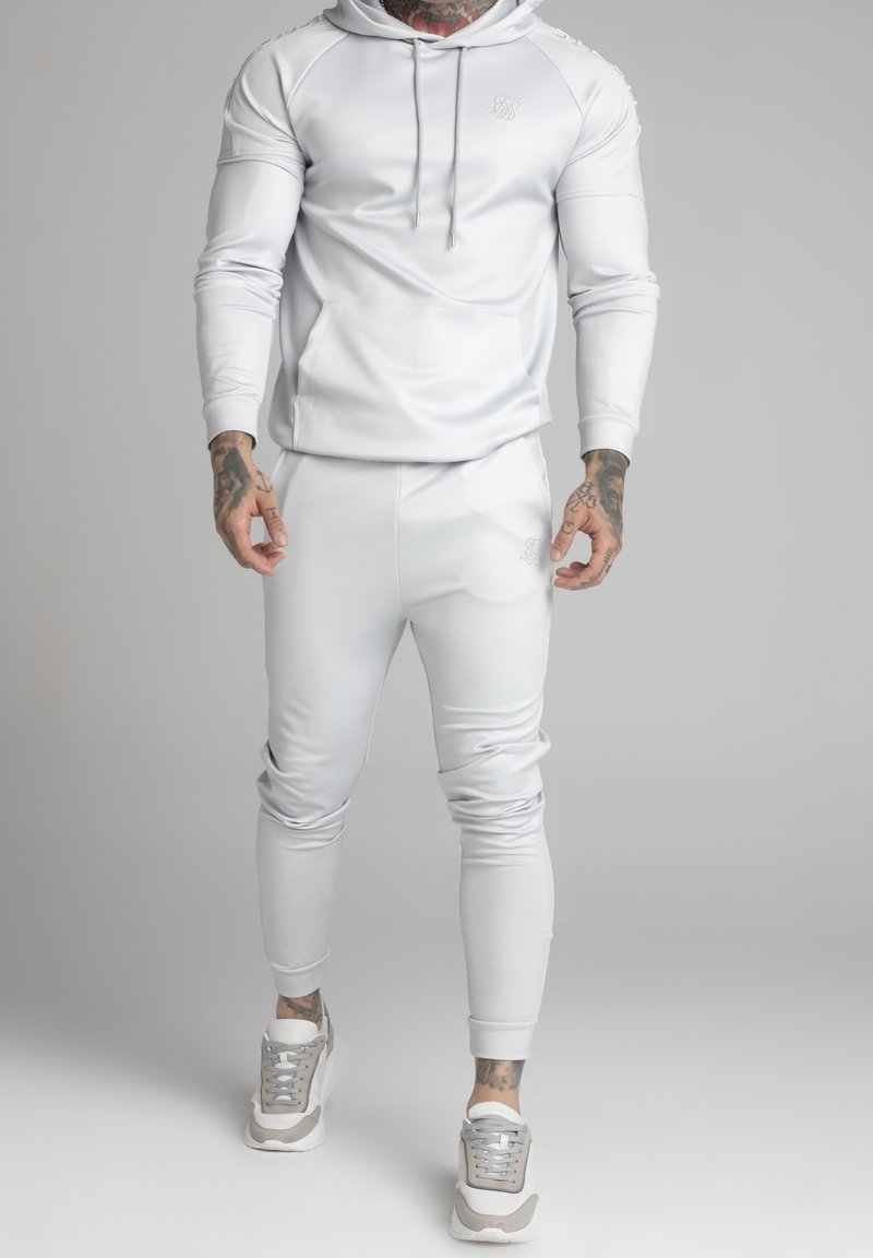 Tuta atletica bianca con cappuccio, tasca frontale e pantaloni aderenti. La texture è liscia; le sneakers sono grigie chiare con accenti beige.