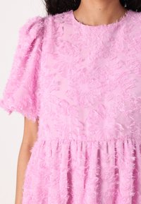 Vestido rosa texturizado com mangas curtas bufantes, apresentando um padrão floral feito de tecido suave e desfiado, e uma cintura franzida.