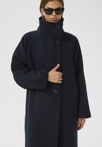 Námořnický kabát s vysokým límcem, oversized střihem a velkými černými knoflíky. Měkká textura a široké rukávy, stylizováno s minimalistickými černými slunečními brýlemi.