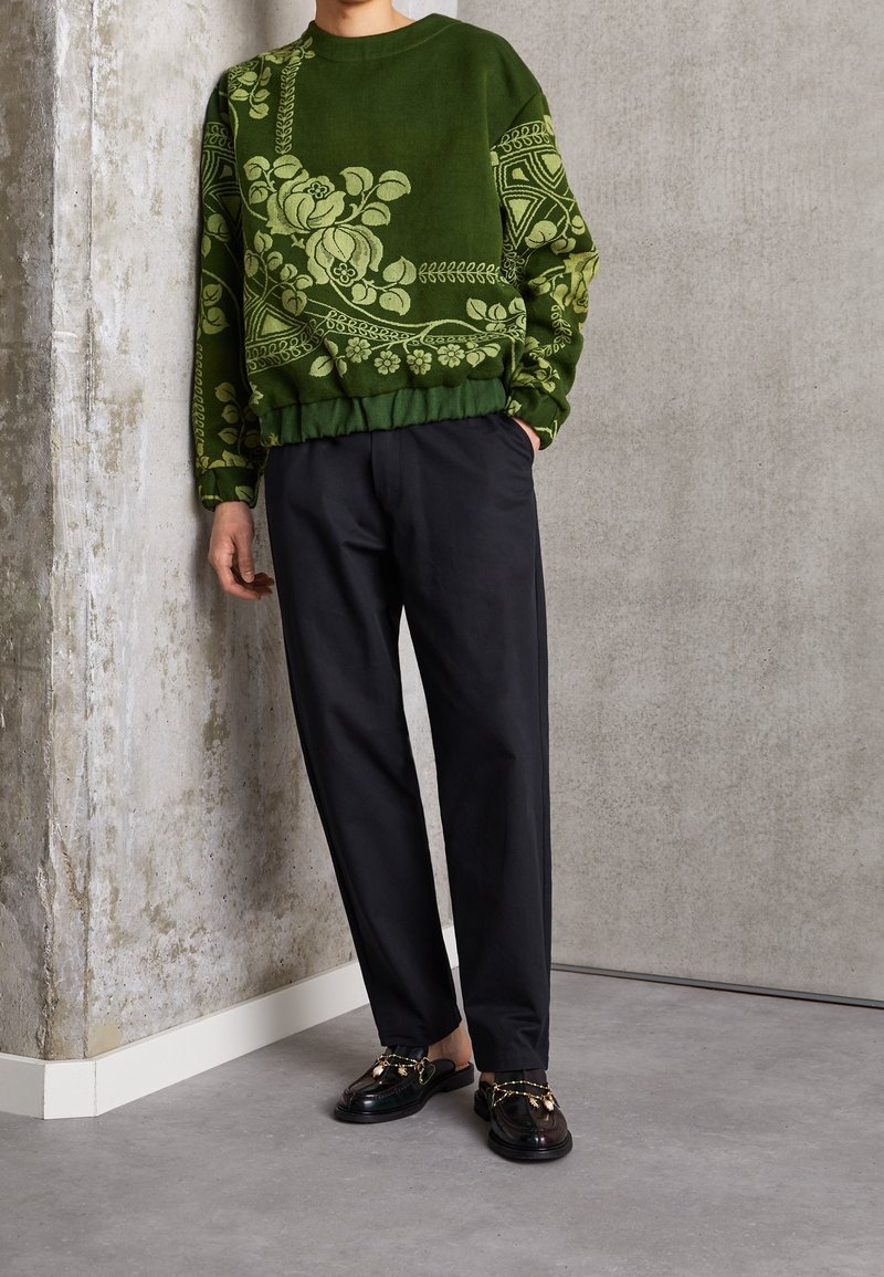 Personne portant un pull vert à motifs floraux, un pantalon noir et des mocassins noirs ornés de chaînes dorées, debout devant un mur texturé.