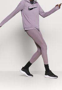 Lila sportliche Langarmoberteile mit einem Halb-Reißverschluss und schwarzem Nike-Logo, kombiniert mit enganliegenden lila Leggings und schwarzen Laufschuhen mit goldenen Akzenten.