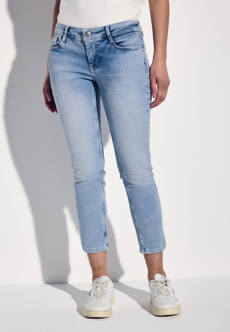 Jeans de mezclilla azul claro con silueta ajustada, dobladillo recortado, cinco bolsillos, trabillas para cinturón y cierre de botón plateado. Combinados con zapatillas blancas.