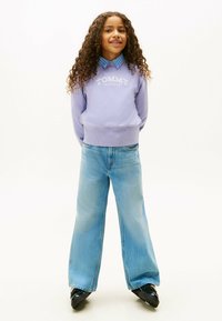 Fioletowa bluza z logo "TOMMY HILFIGER", jasnoniebieska koszula z guzikiem, wysokie niebieskie dżinsy o szerokich nogawkach i czarne buty.