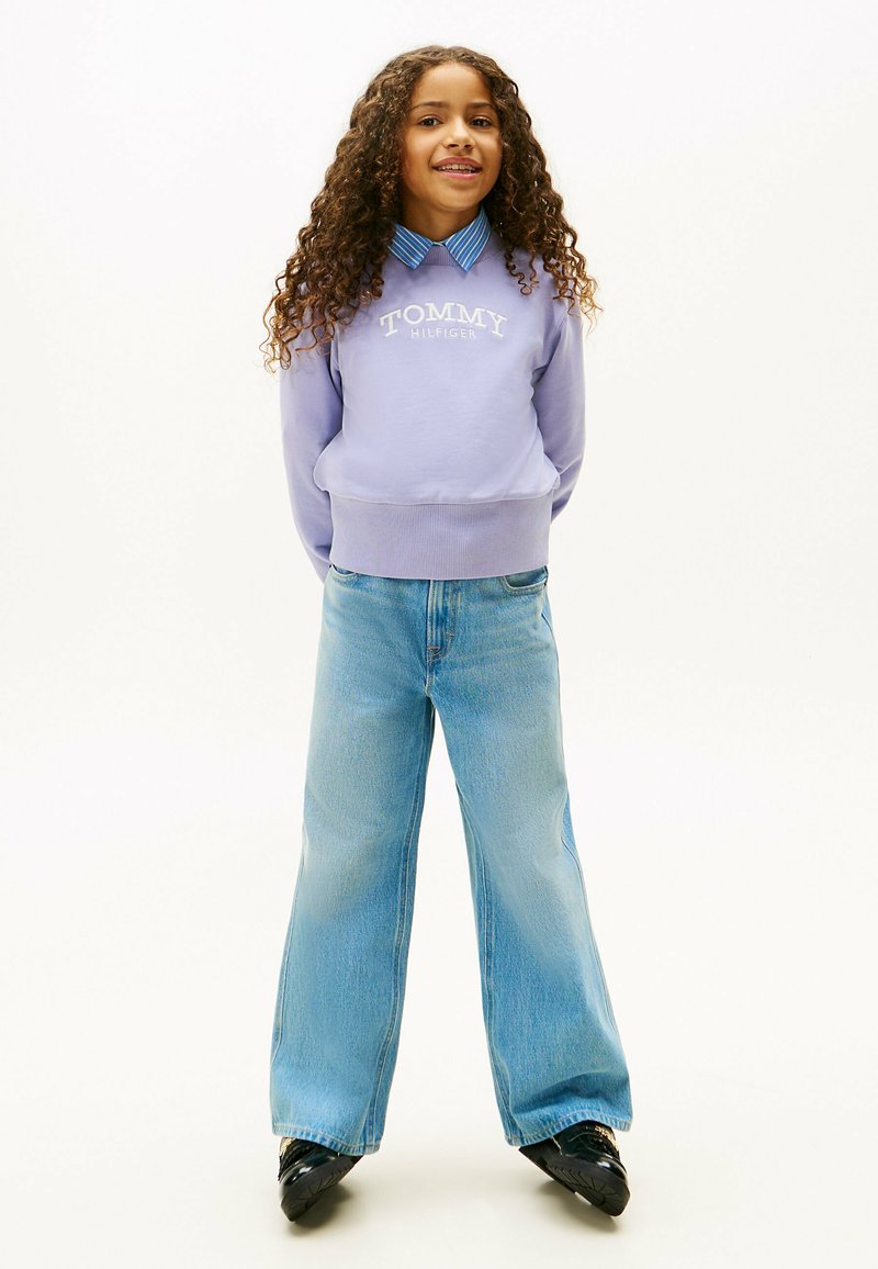 Fioletowa bluza z logo "TOMMY HILFIGER", jasnoniebieska koszula z guzikiem, wysokie niebieskie dżinsy o szerokich nogawkach i czarne buty.