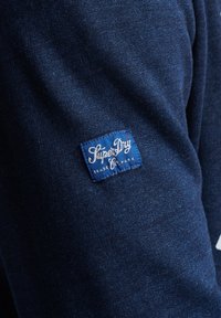 Felpa di colore blu navy con una superficie texturizzata, dotata di un'etichetta blu con testo bianco che recita "SuperDry Trade Mark."