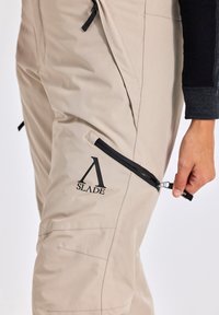 Beige buitenbroek met zwarte ritsen en accenten. Voorzien van een ritszak op de dij en een logo nabij de zak. Glad, duurzaam materiaal.