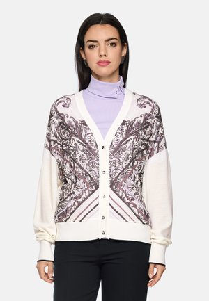 Frau trägt einen weißen, geknöpften Cardigan mit lila und schwarzen Paisley-Mustern über einem lavendelfarbenen Rollkragenpullover und schwarzen Hosen.