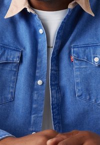 Camicia di jeans blu con colletto beige, dotata di due tasche sul petto e bottoni bianchi. Indossata sopra una maglietta bianca.